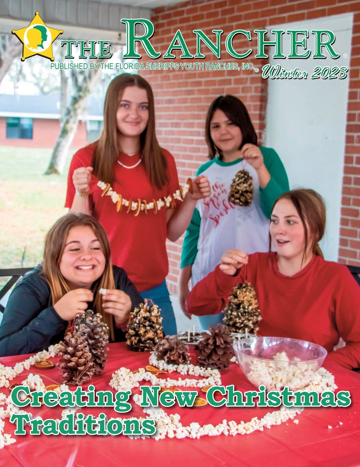 This is the cover of the 2023 Winter edition of the Rancher magazine. It depicts three girls having fun making decorations for a Christmas tree, They are stringing popcorn together and decorating pinecones.